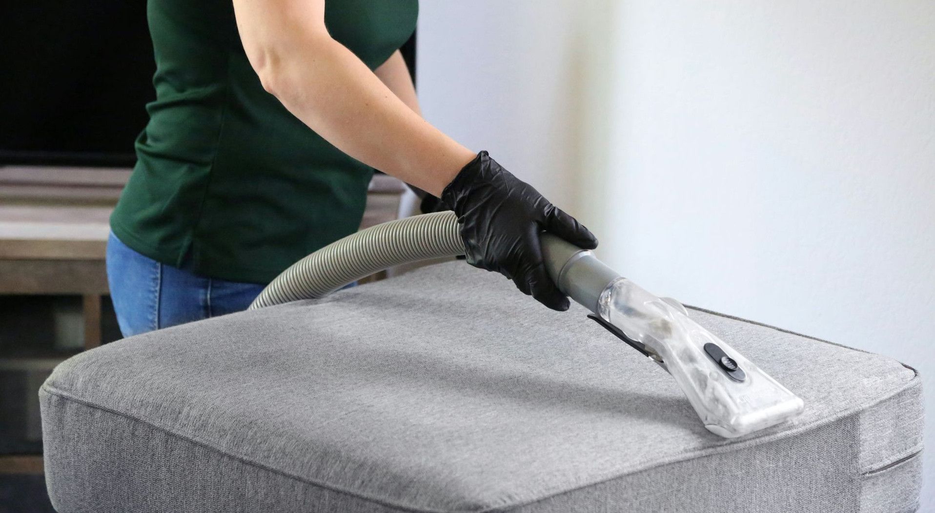 a woman in a green shirt and black gloves vacuuming a gray ottoman