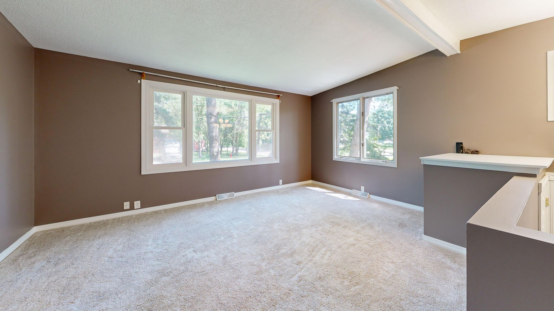 Large empty living room with neutral walls and carpet, natural light through windows.