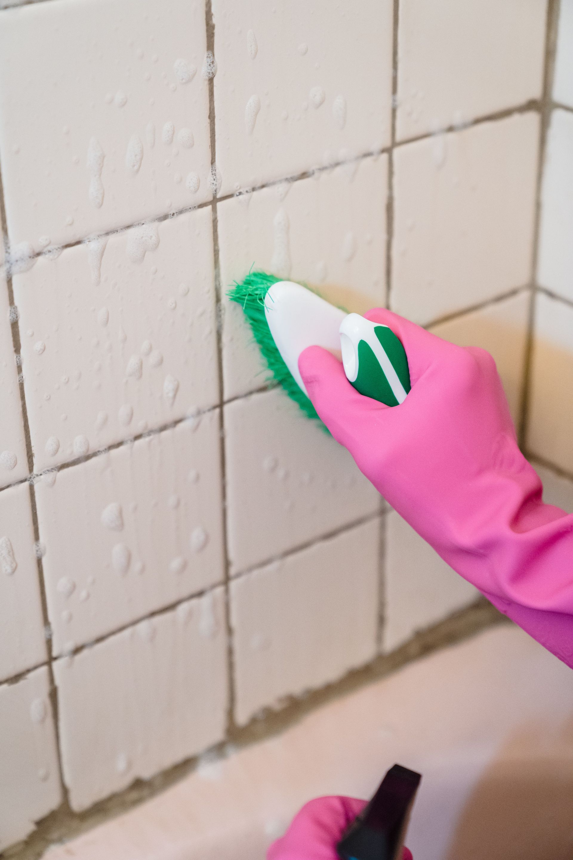 A hand in pink gloves scrubs bathroom tiles with a green brush to remove grime.
